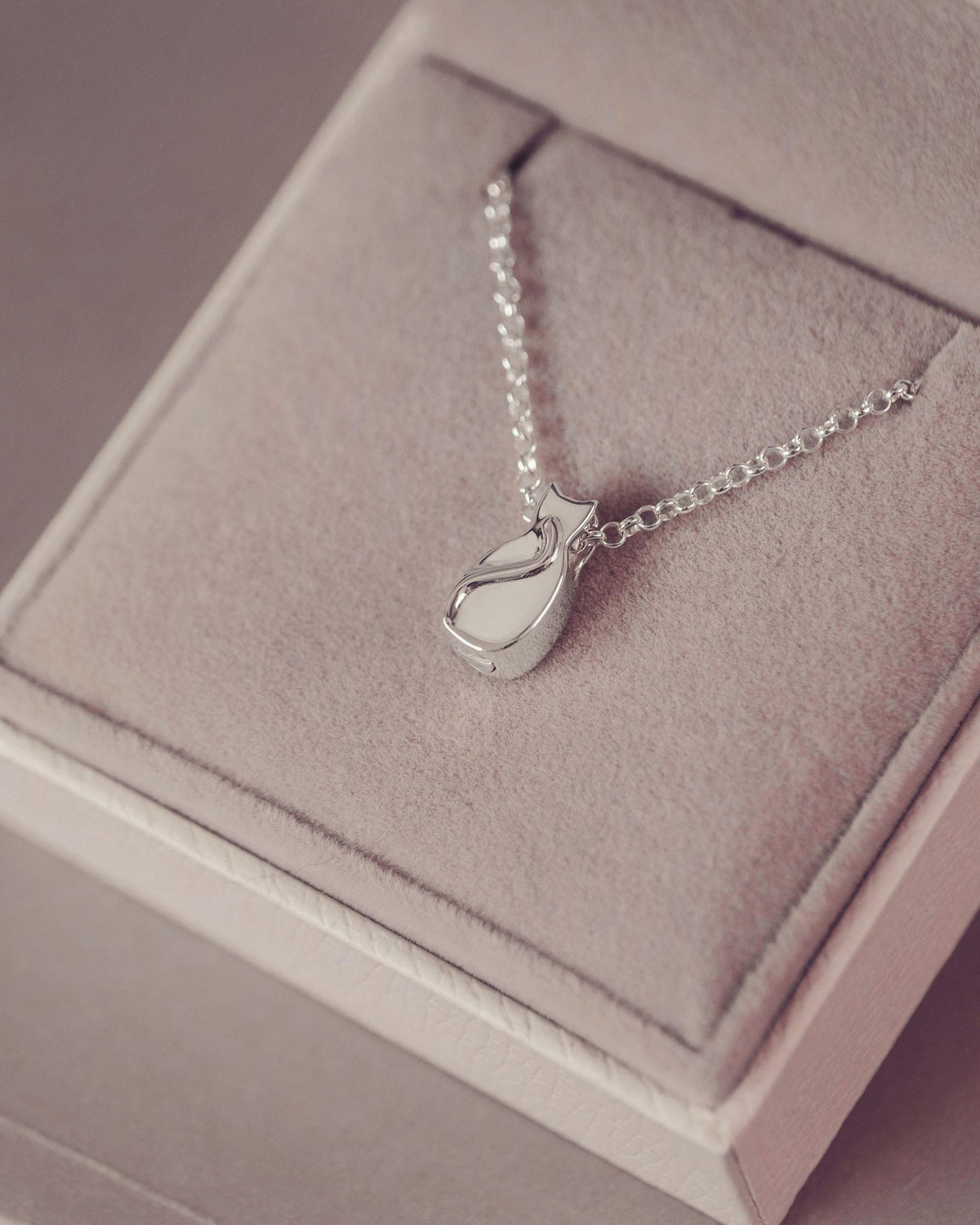 A silver pendant necklace with a small stylized cat silhouette, displayed on a soft pink jewelry box.