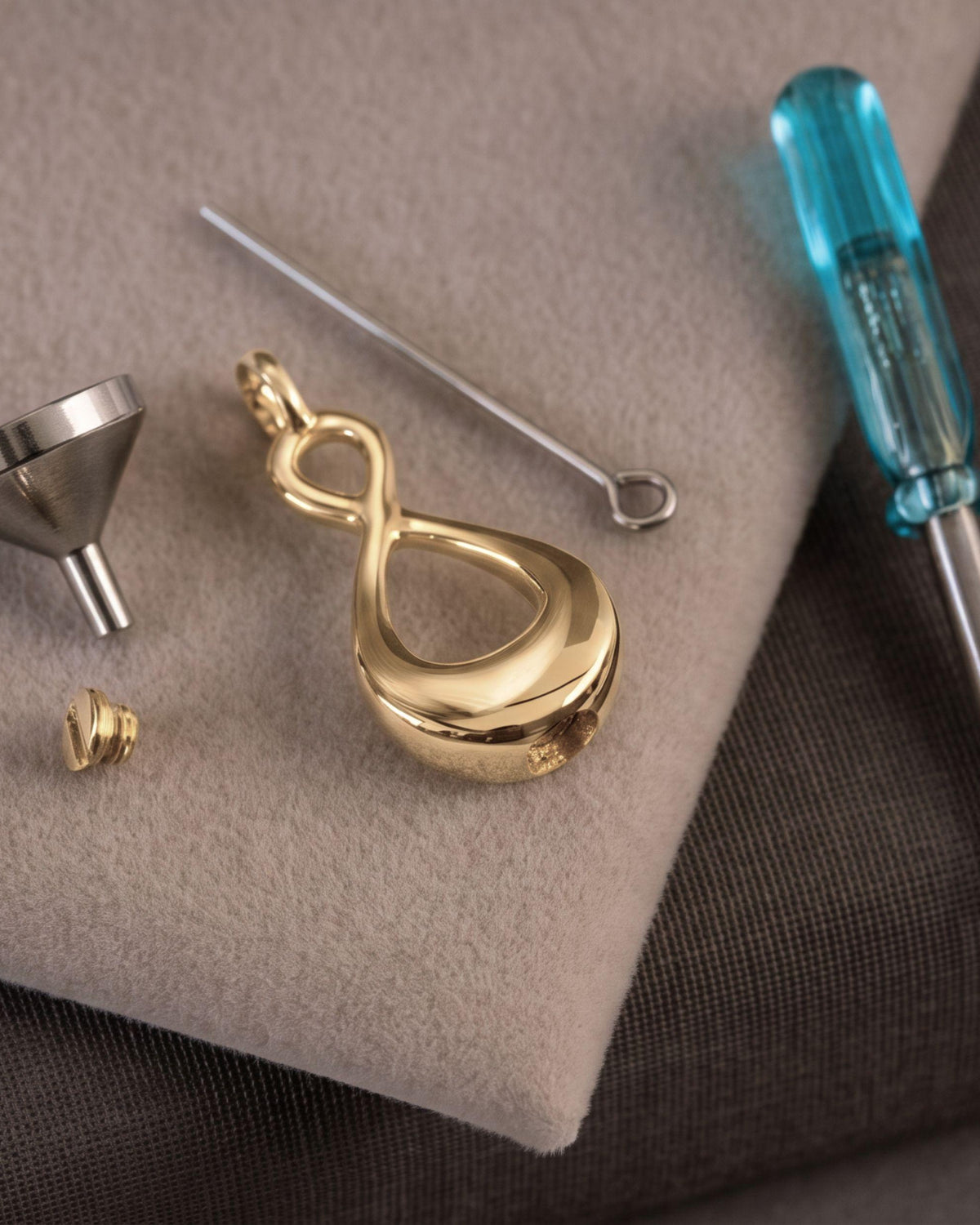 A gold infinity-shaped pendant charm displayed with its silver metal funnel, small gold cap, and a blue-handled screwdriver on a beige textured mat.