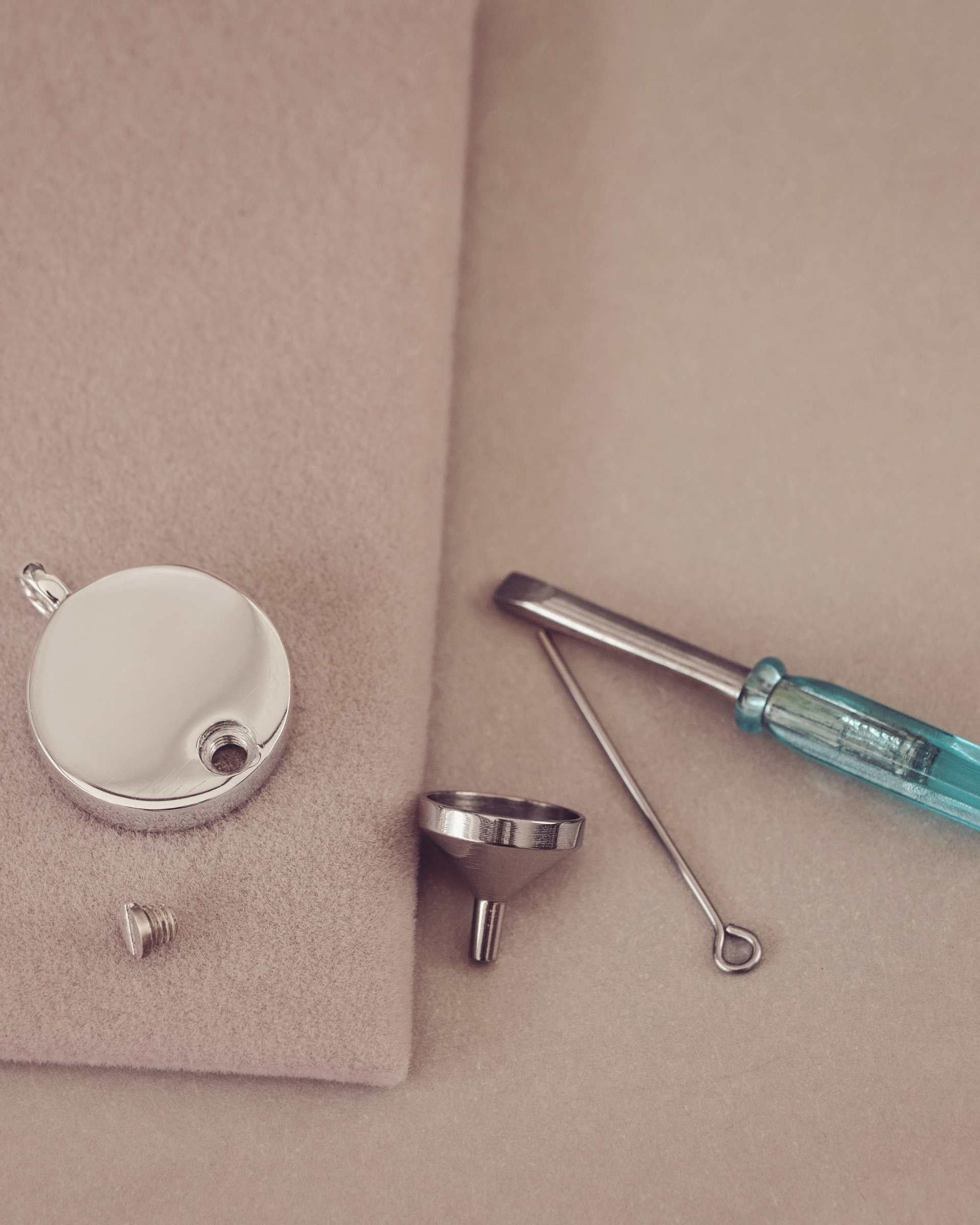 Jewellery tools including a silver pendant, screwdriver, funnel, and small metal component on a beige surface.