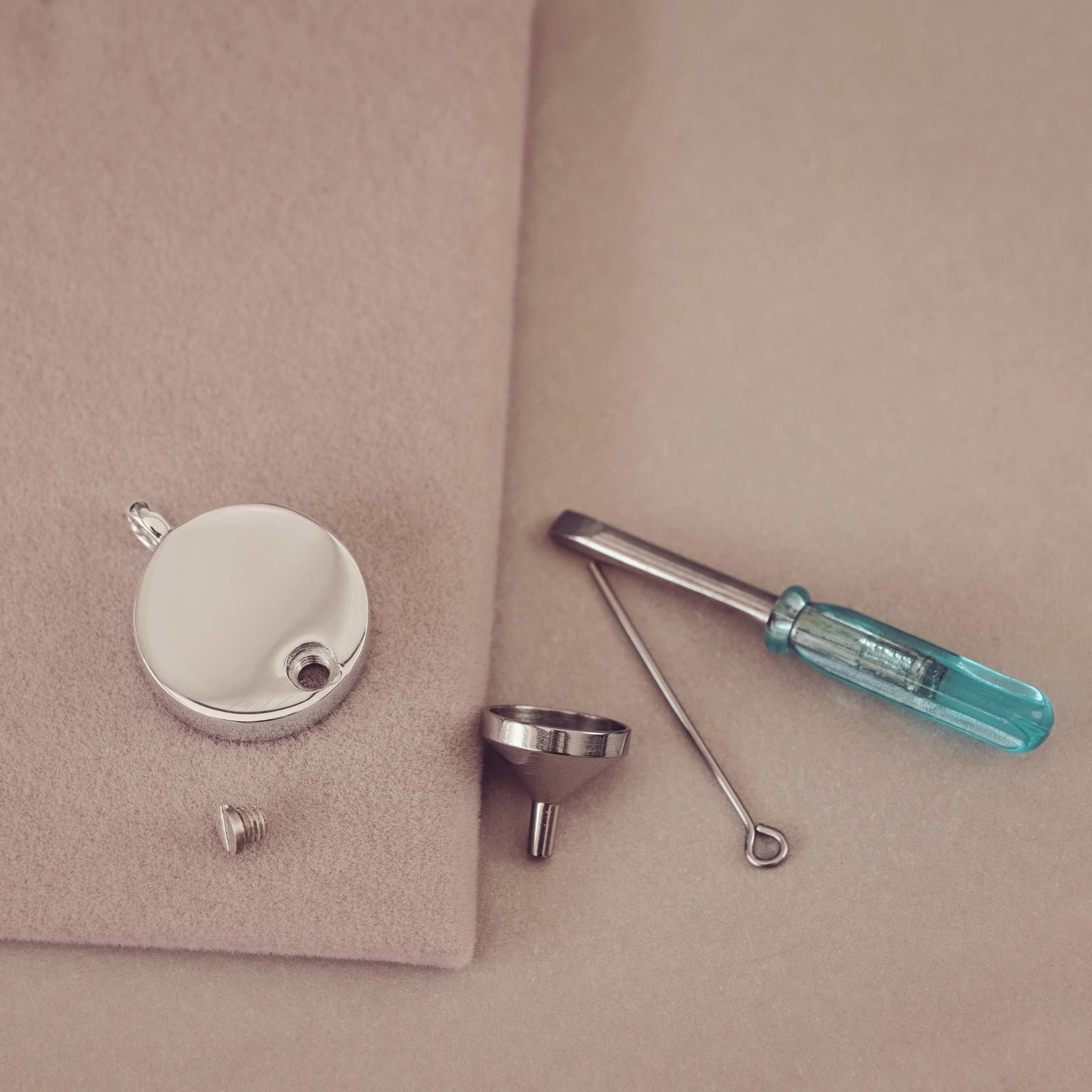 Jewellery tools including a silver pendant, screwdriver, funnel, and small metal component on a beige surface.