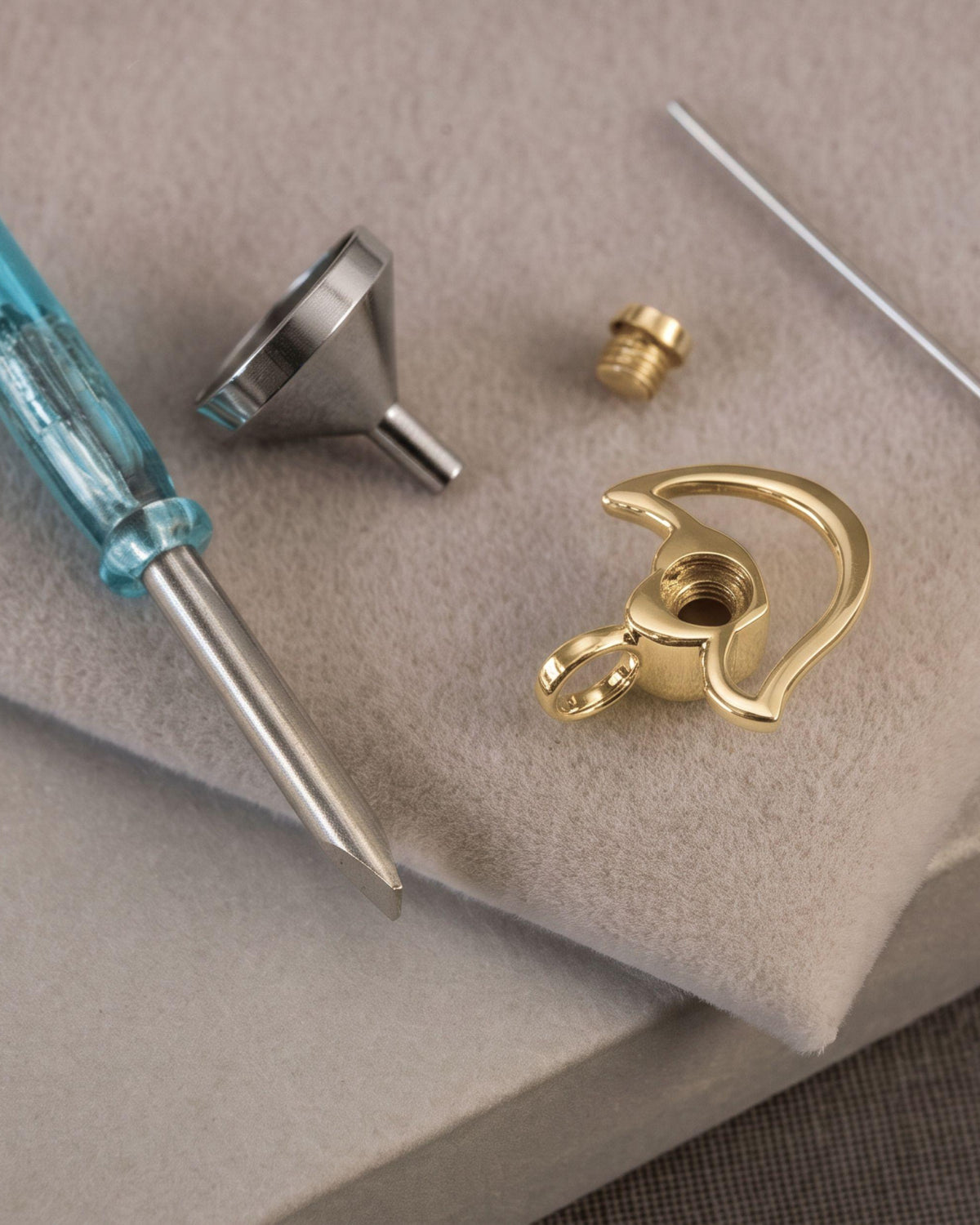 Close-up of a small tools on a beige textured surface, including a blue-handled flathead screwdriver, a metal funnel, a small brass screw, a curved gold pendant piece, and a thin metal pin.