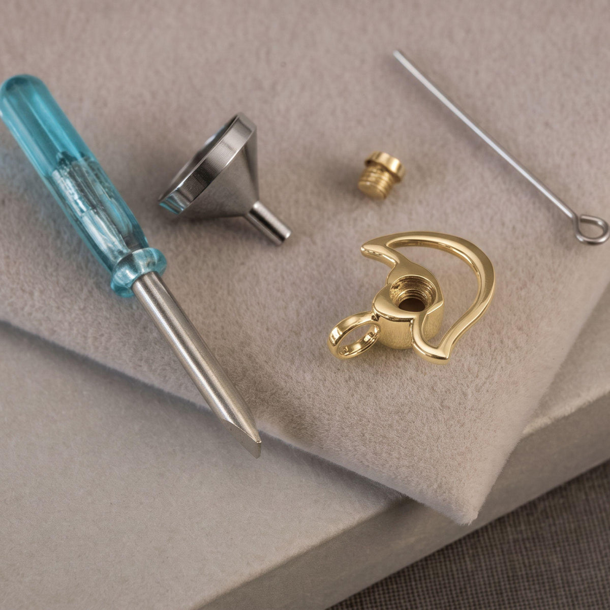 Close-up of a small tools on a beige textured surface, including a blue-handled flathead screwdriver, a metal funnel, a small brass screw, a curved gold pendant piece, and a thin metal pin.