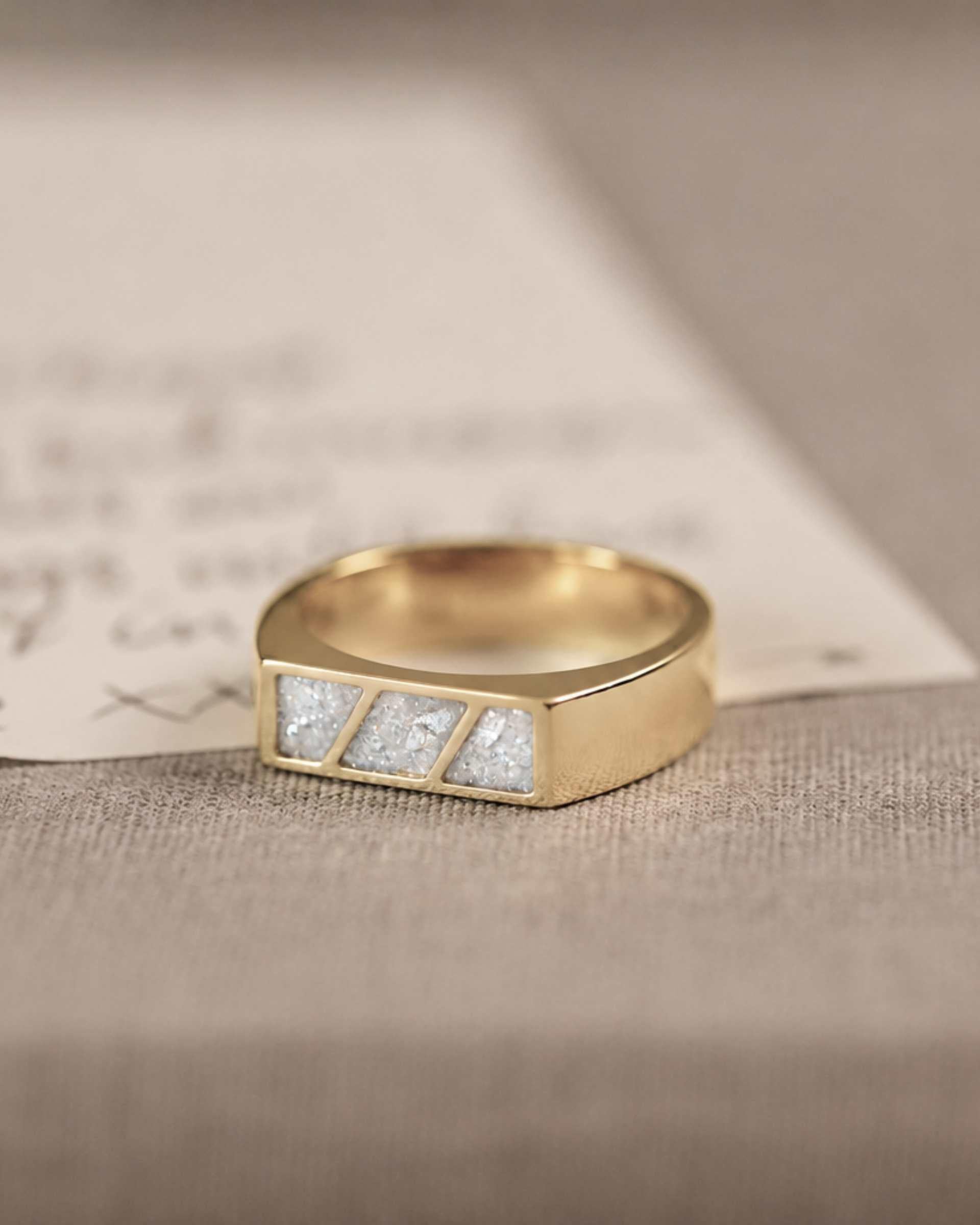 A gold ring with 3 sections of white resin across the top. Sitting on a hand written letter and textured background