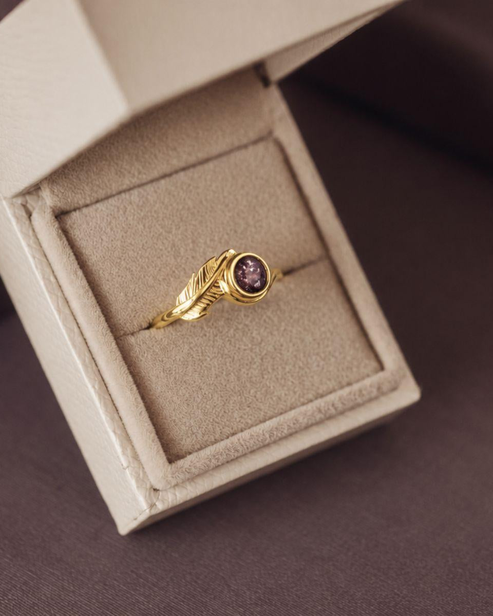 A gold ring with a leaf-shaped band and a round purple gemstone, displayed inside an open beige jewelry box on a dark brown surface.