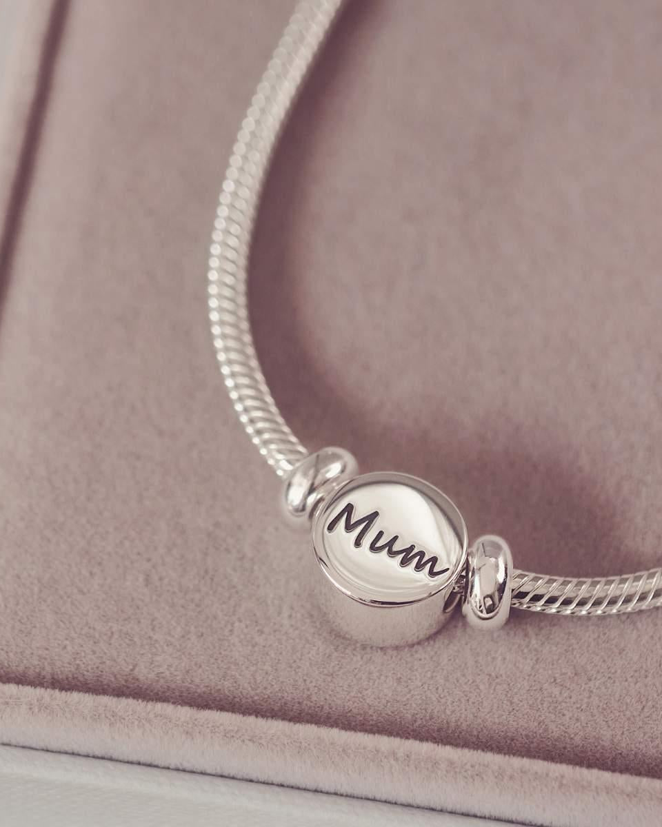 Silver bracelet with a round charm inscribed with 'Mum' on a pink surface