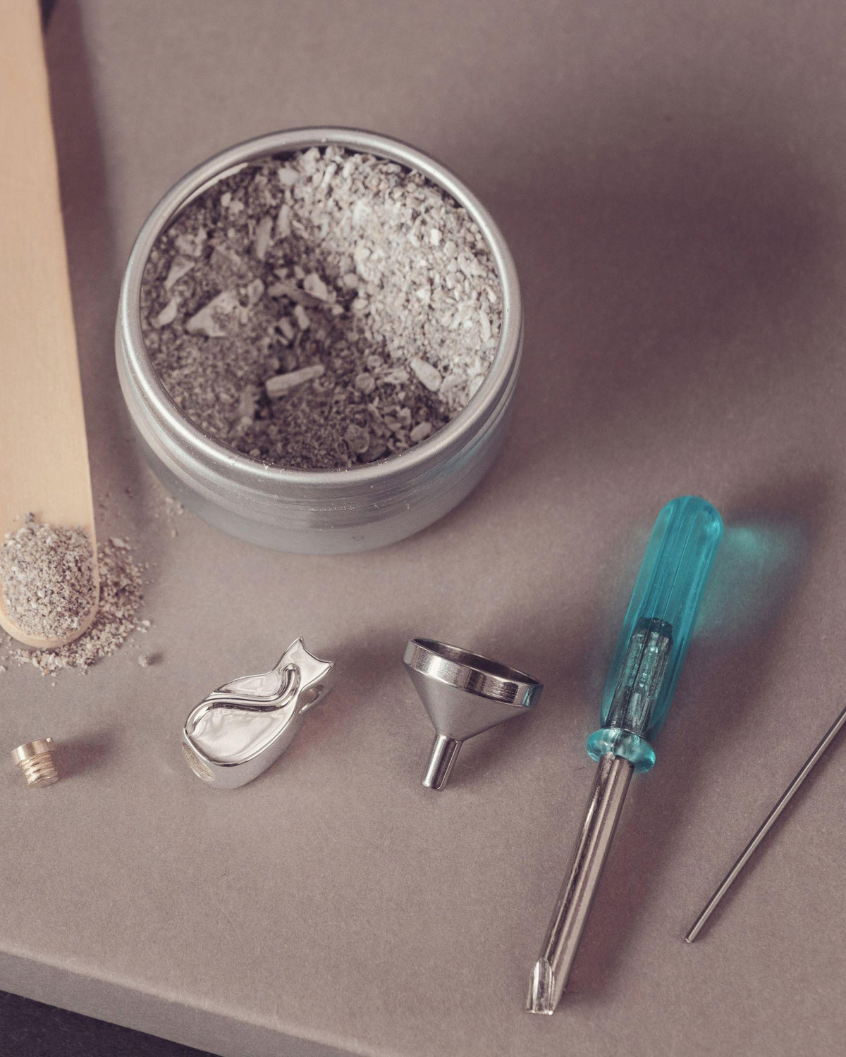 A metal tin filled with ashes, arranged on a taupe surface with a wooden spatula holding some ash, a small metal screw, a silver funnel, ametal piece, and a turquoise-handled tool.