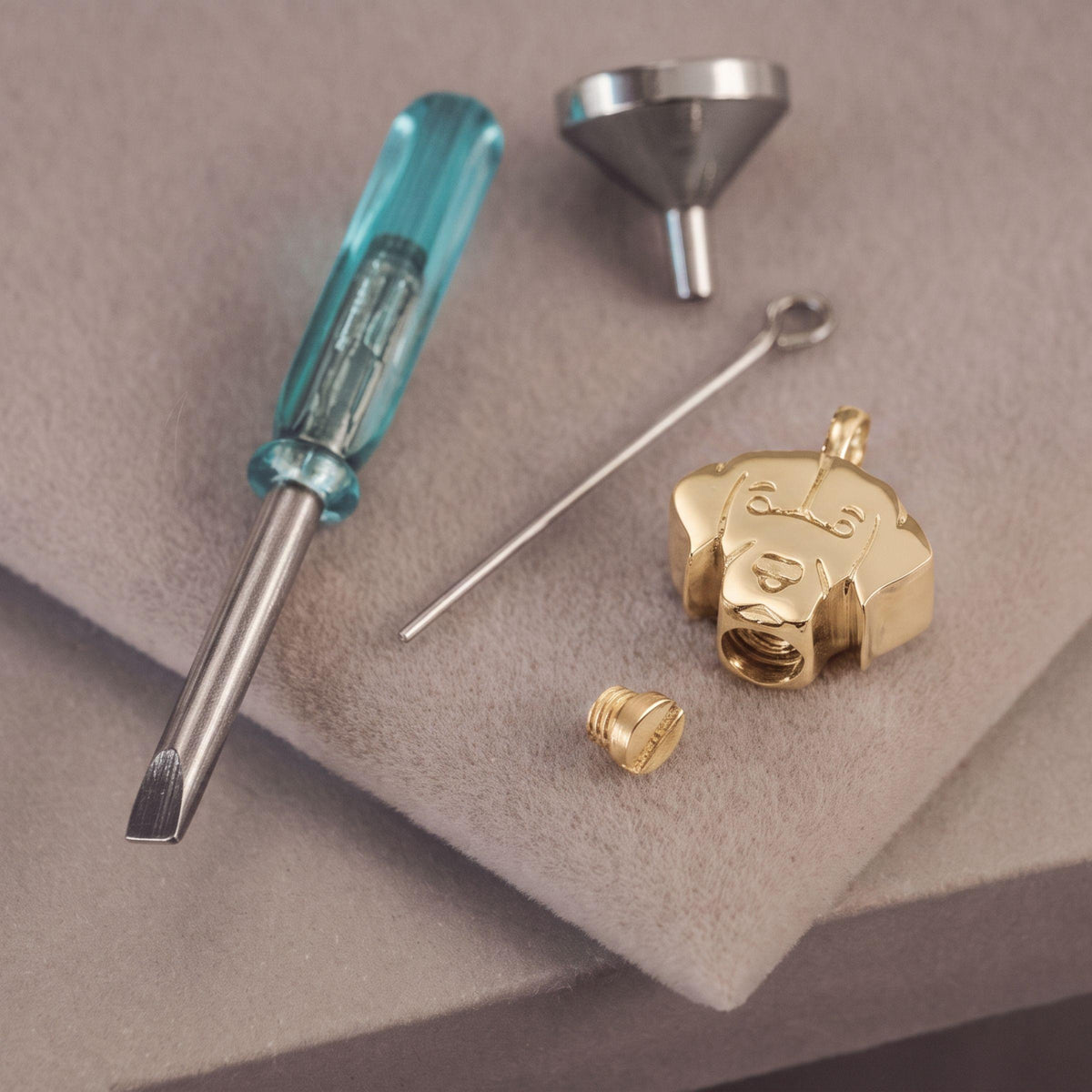 A gold labrador dog pendant charm shown disassembled with a small screwdriver, a funnel, and a pin on a textured gray background.