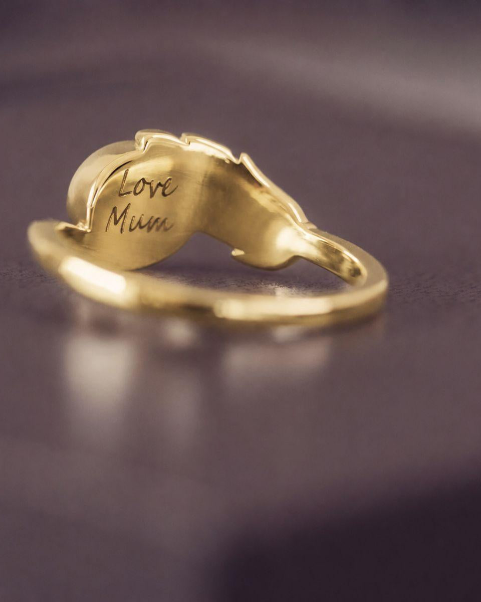 A gold ring shaped like a mother's hand holding a heart, with 'Love Mum' engraved on the heart, resting on a dark purple surface.