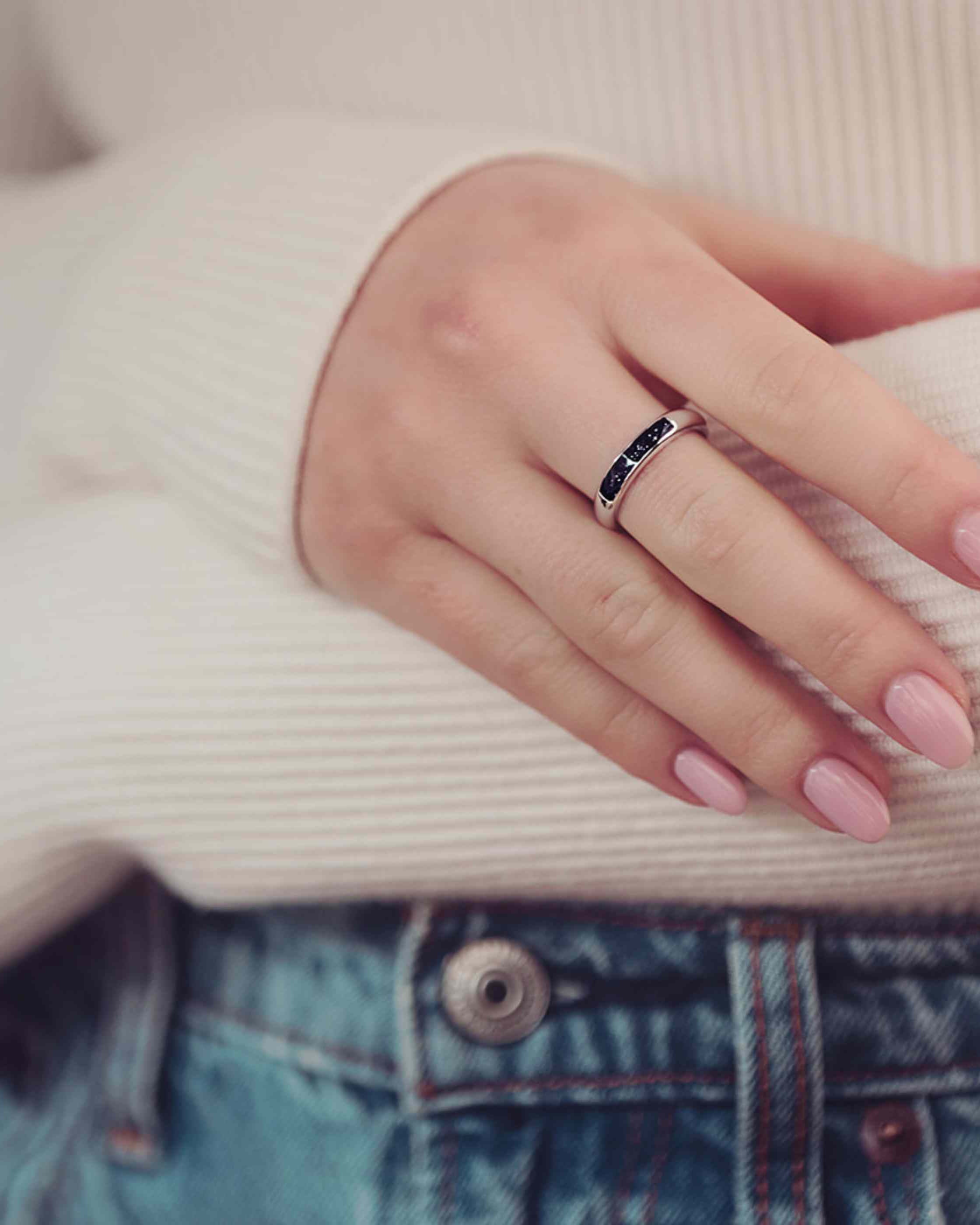 close up a womens hand Womens Cremation Ashes Ring Band, filled with ashes and black crushed crystal. The lady has pink painted nails and is wearing a white jumper