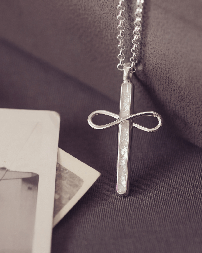 Silver cross necklace with infinity symbol on a textured surface with a black and white photo.