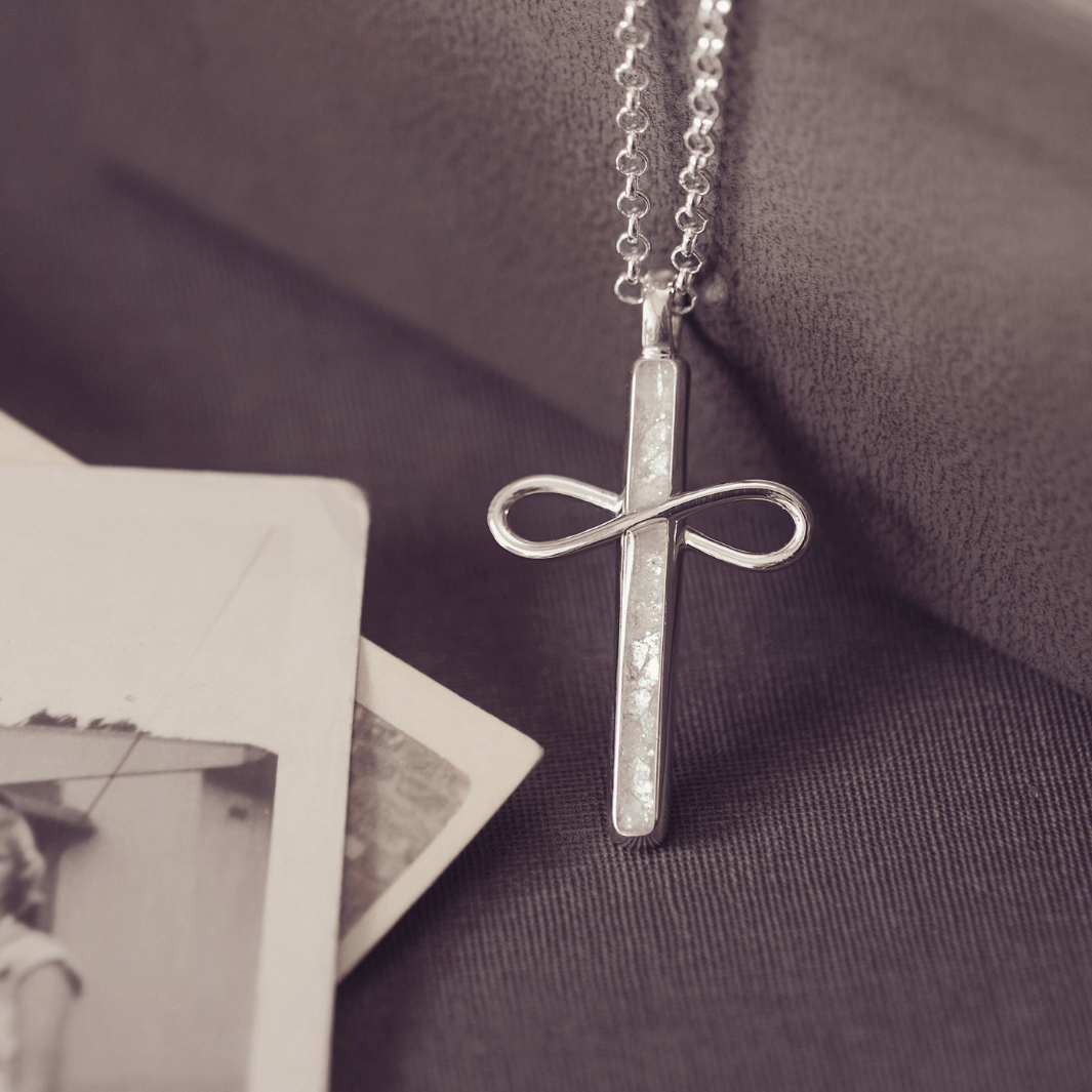 Silver cross necklace with infinity symbol on a textured surface with a black and white photo.