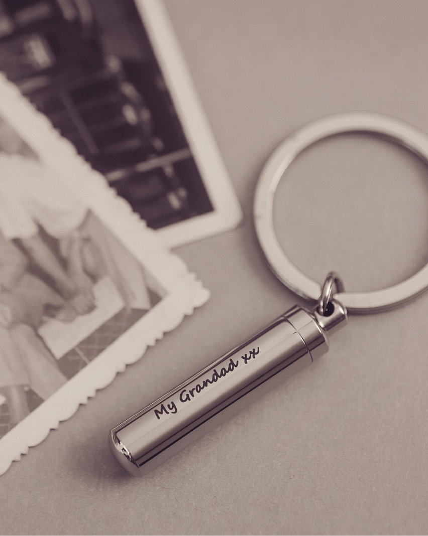 Cylindrical keyring which unscrews to hold cremation ashes or hair, engraved with the message "My Grandad xx" on one line, on a light grey background with vintage photos in the background.