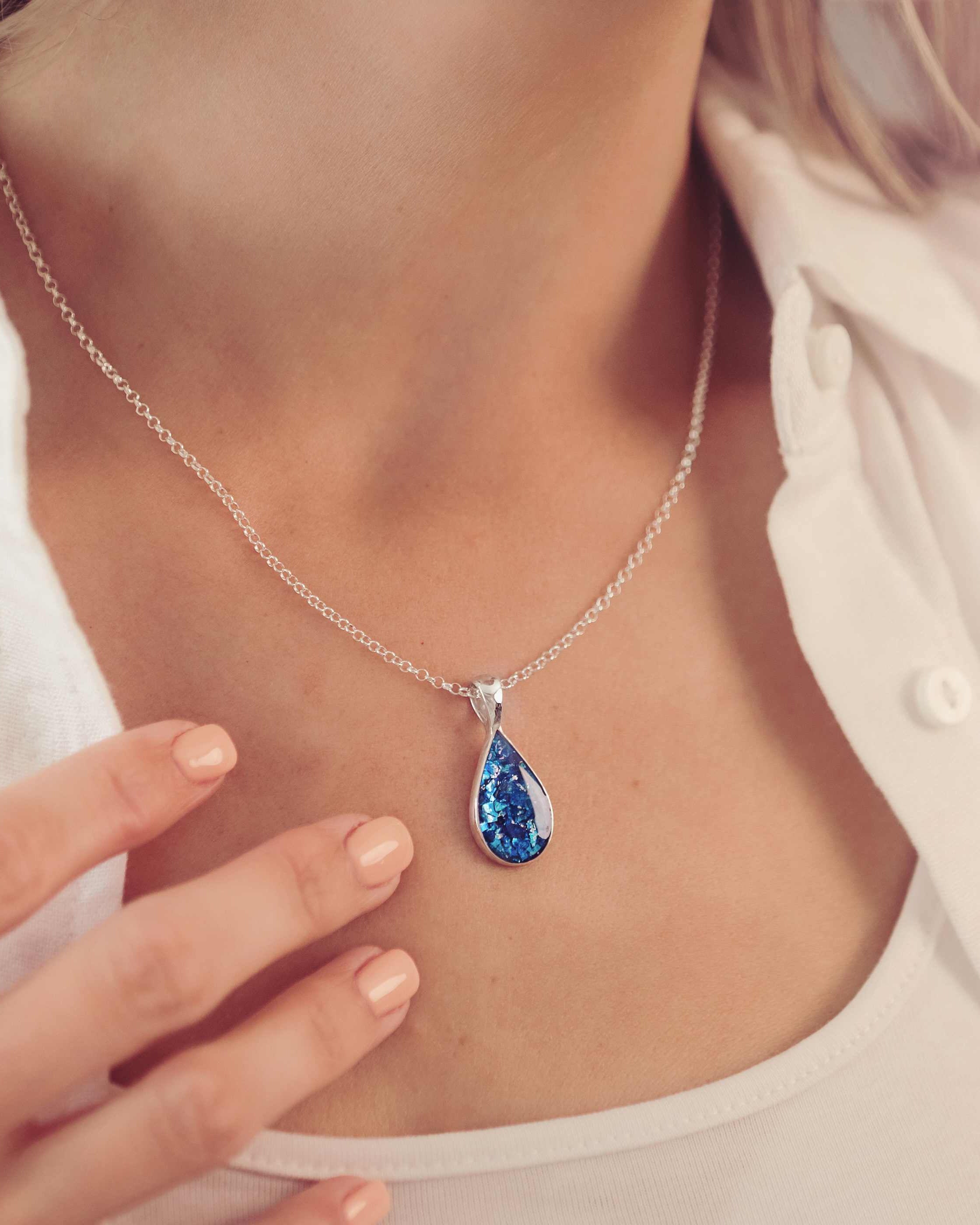 lady wearing a blue teardrop shaped ashes necklace filled with cremation ashes, she is wearing a white shirt and has beige painted nails