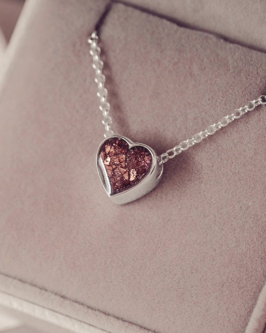 A silver heart pendant necklace with a textured, glittery brown center, resting on a dusty pink velvet jewelry box.