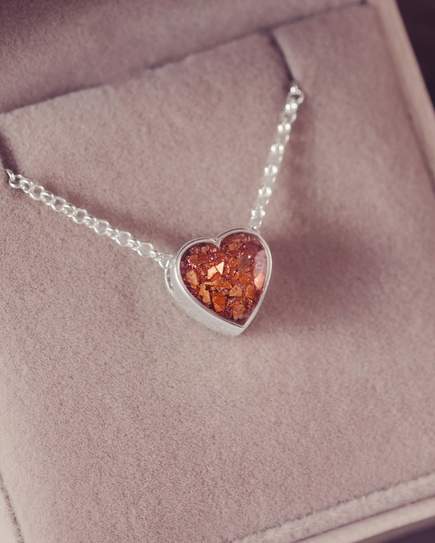 A silver heart pendant necklace with amber-colored inclusions, displayed inside a soft pink jewelry box.