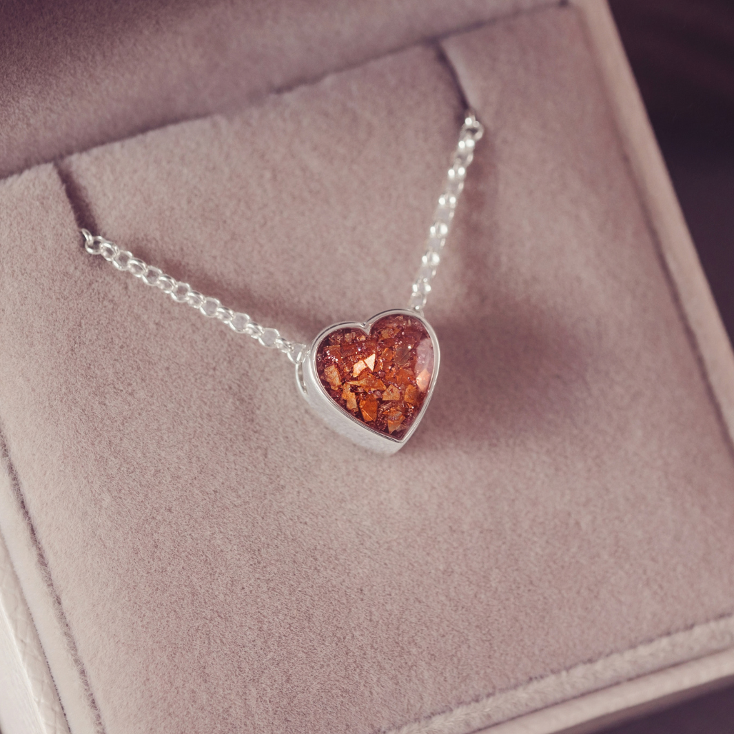 A silver heart pendant necklace with amber-colored inclusions, displayed inside a soft pink jewelry box.