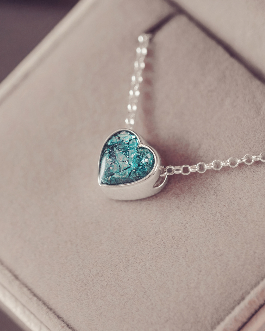 A silver heart-shaped pendant necklace with a textured blue and green mosaic glass center, resting on a beige jewelry box.