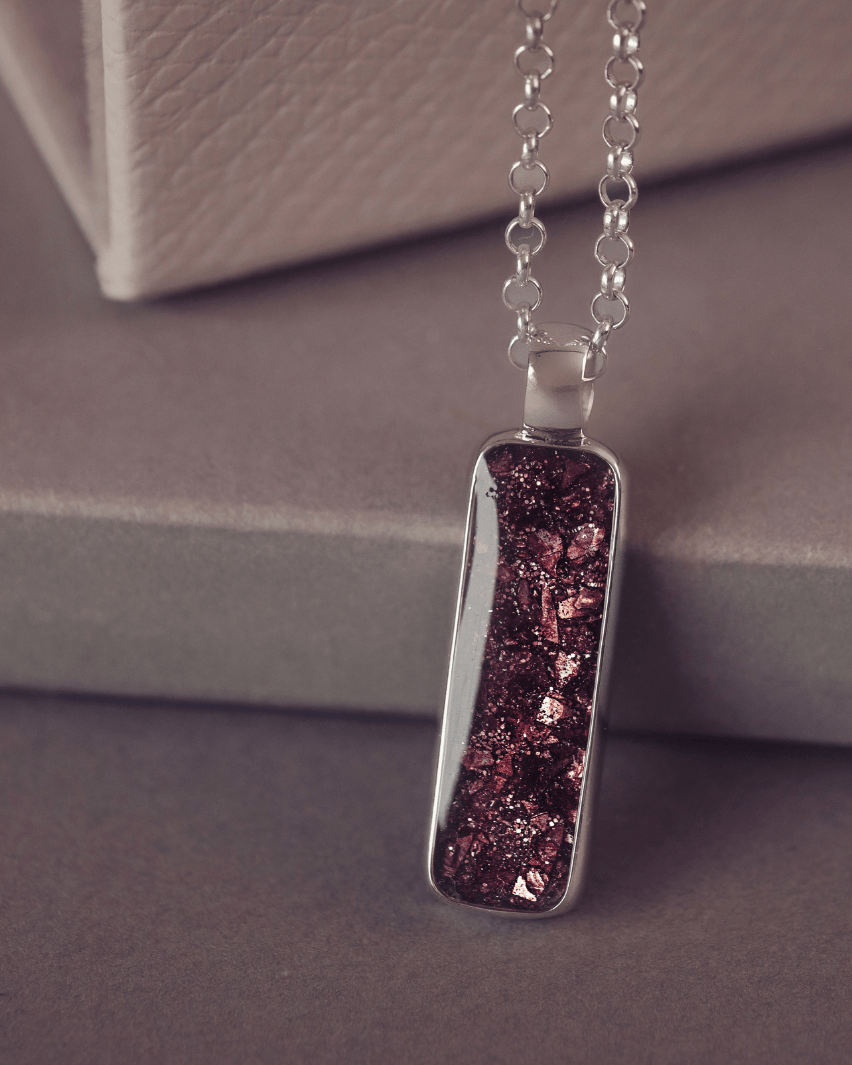 A sophisticated silver necklace featuring a rectangular pendant filled with shimmering brown crystal and resin, delicately layered with cremation ashes, showcased against a dark background, on a silver belcher chain.