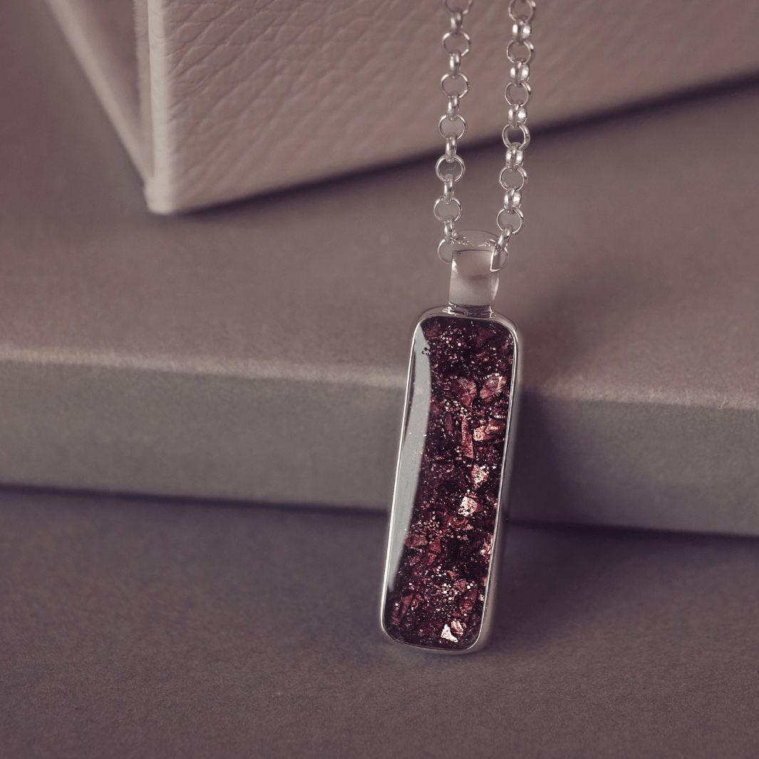 A sophisticated silver necklace featuring a rectangular pendant filled with shimmering brown crystal and resin, delicately layered with cremation ashes, showcased against a dark background, on a silver belcher chain.