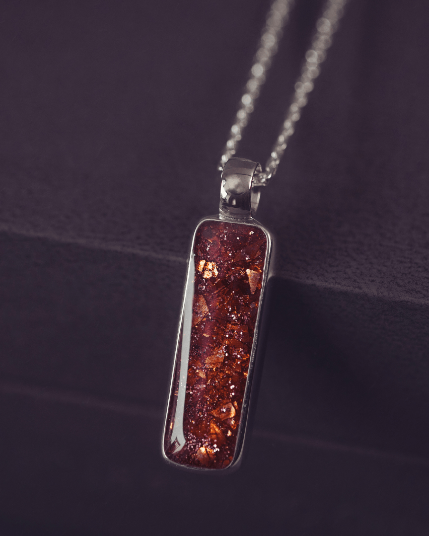 A sophisticated silver necklace featuring a rectangular pendant filled with shimmering orange crystal and resin, delicately layered with cremation ashes, showcased against a dark background, on a silver belcher chain.