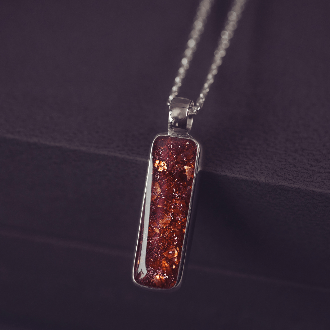 A sophisticated silver necklace featuring a rectangular pendant filled with shimmering orange crystal and resin, delicately layered with cremation ashes, showcased against a dark background, on a silver belcher chain.