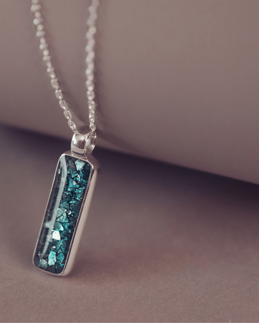 A sophisticated silver necklace featuring a rectangular pendant filled with shimmering turquoise crystal and resin, delicately layered with cremation ashes, showcased against a dark background, on a silver belcher chain.