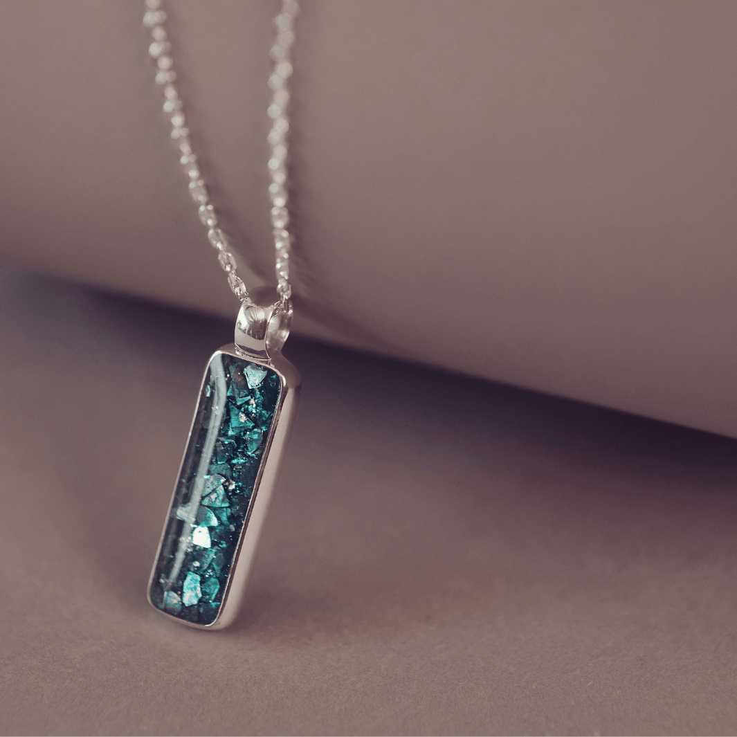 A sophisticated silver necklace featuring a rectangular pendant filled with shimmering turquoise crystal and resin, delicately layered with cremation ashes, showcased against a dark background, on a silver belcher chain.