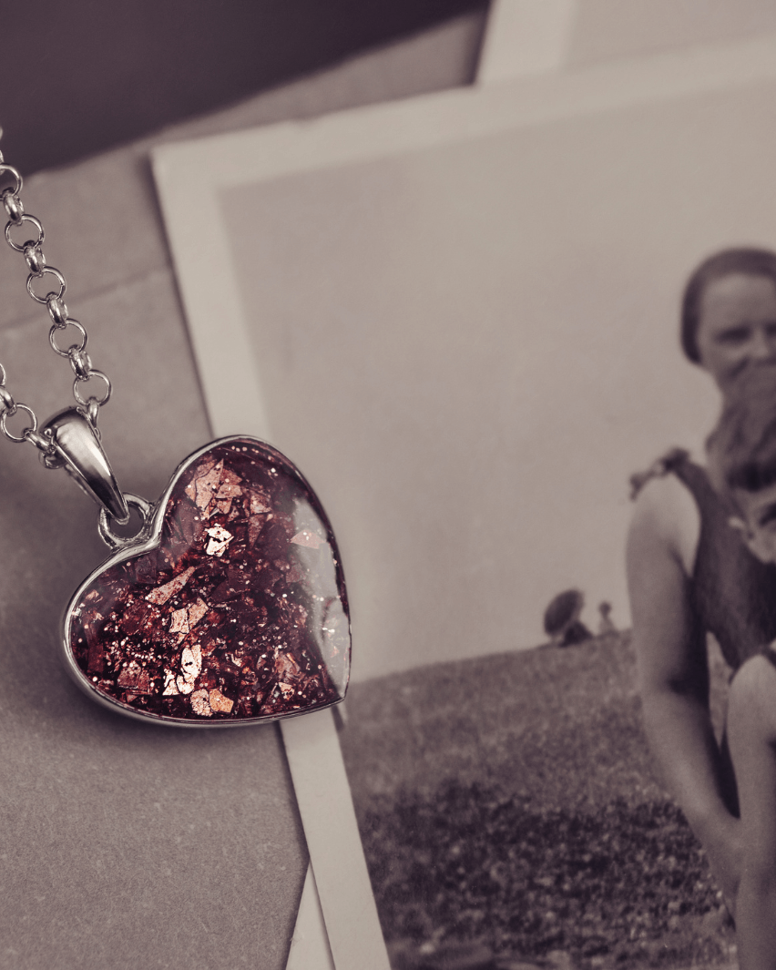 Sterling silver heart necklace with a central section filled with memorial ashes and brown crushed crystals, displayed on a beige fabric background next to a black and white photograph