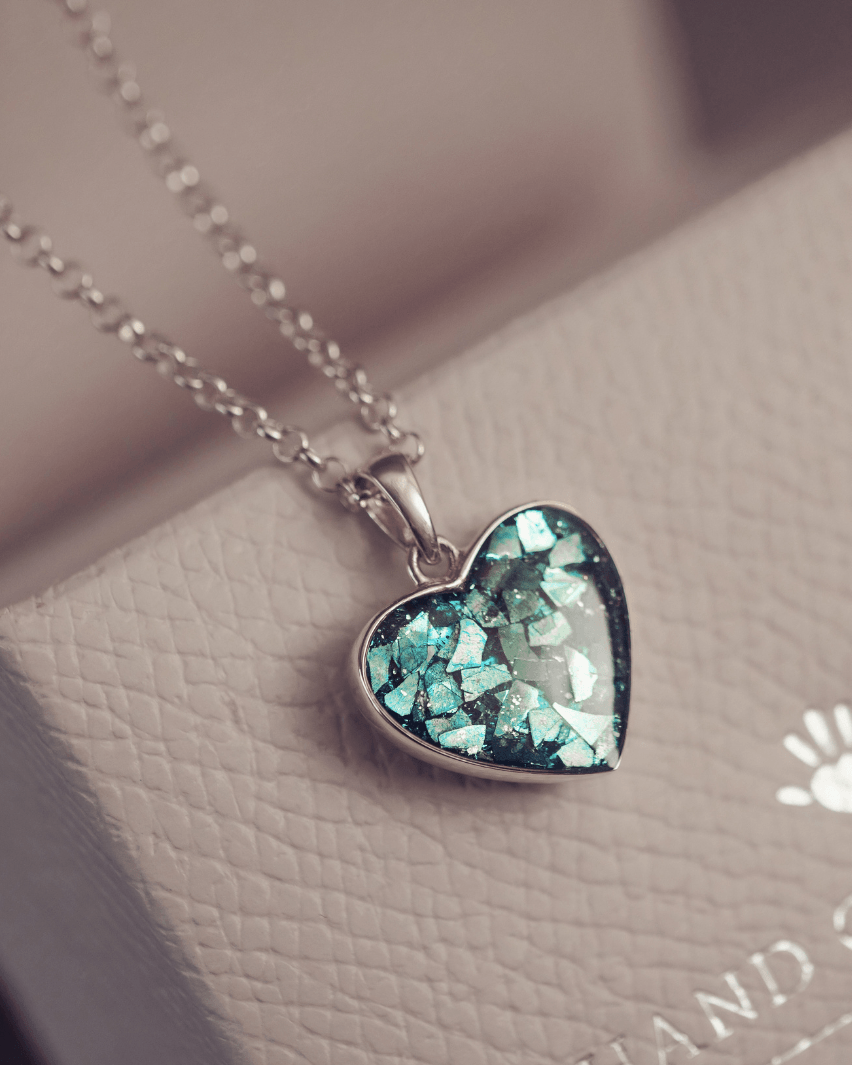 Sterling silver heart necklace with a central section filled with memorial ashes and turquoise crushed crystals, displayed on a grey textured jewellery box