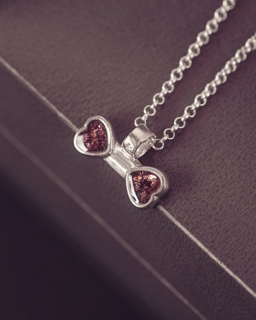 A silver chain necklace with a bow-shaped pendant featuring two heart-shaped brown gemstone inlays, displayed on a dark textured surface.