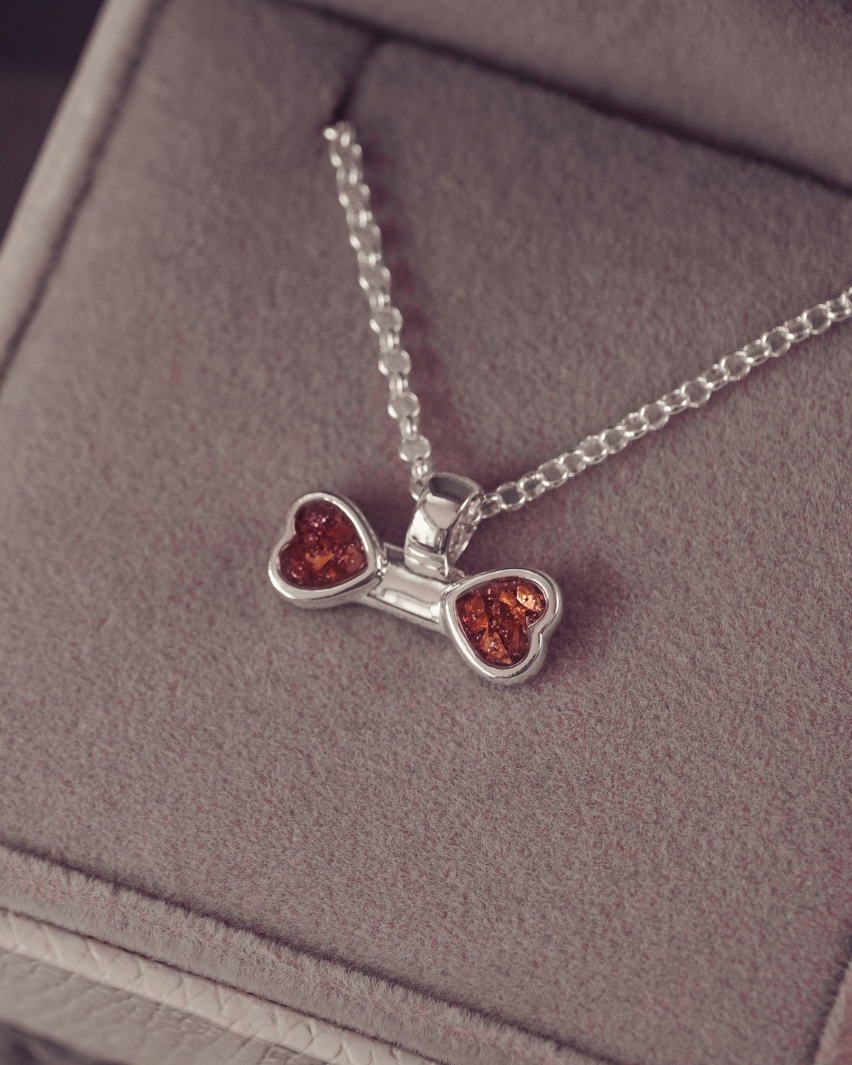 A silver necklace with a bow-shaped pendant featuring two amber heart inlays, displayed inside a gray jewelry box.