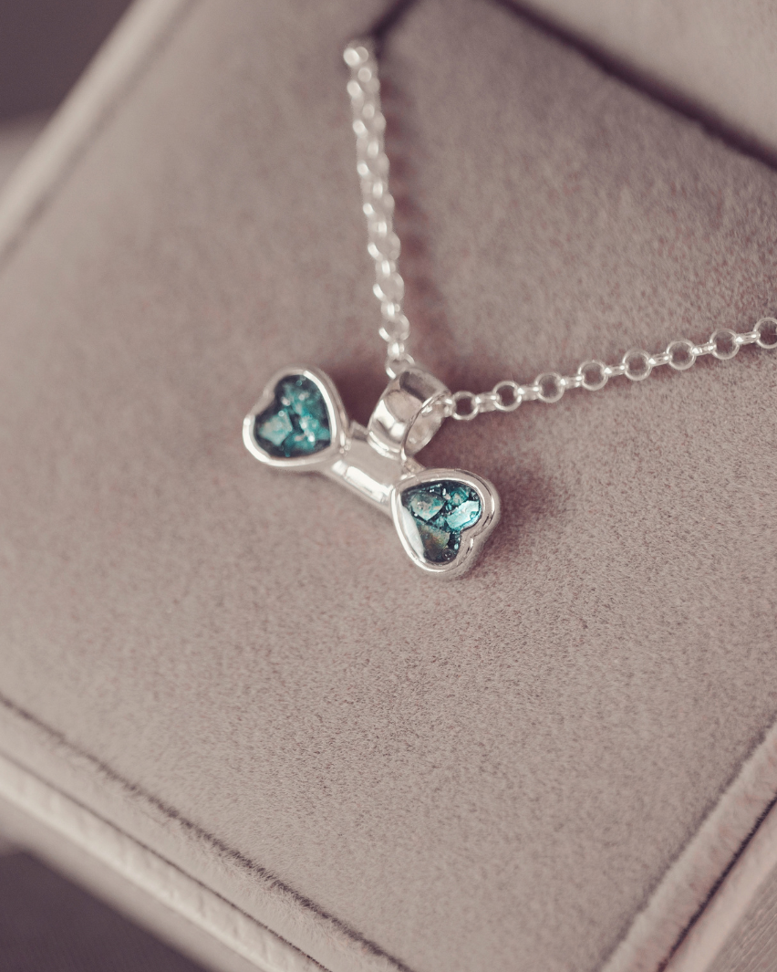 A silver necklace with a bone-shaped pendant featuring two heart-shaped blue-green crystal inlays, resting on a beige jewelry box.