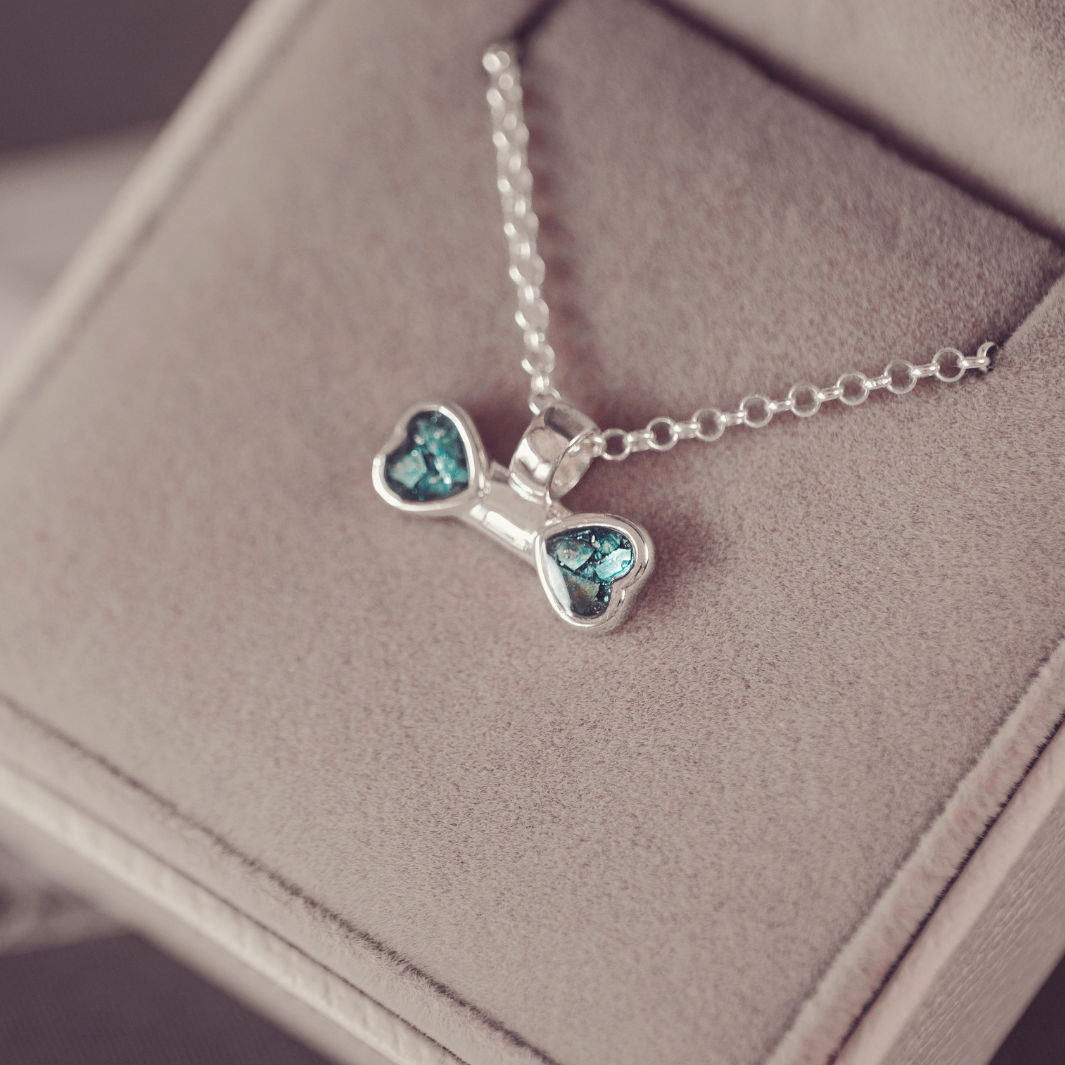 A silver necklace with a bone-shaped pendant featuring two heart-shaped blue-green crystal inlays, resting on a beige jewelry box.
