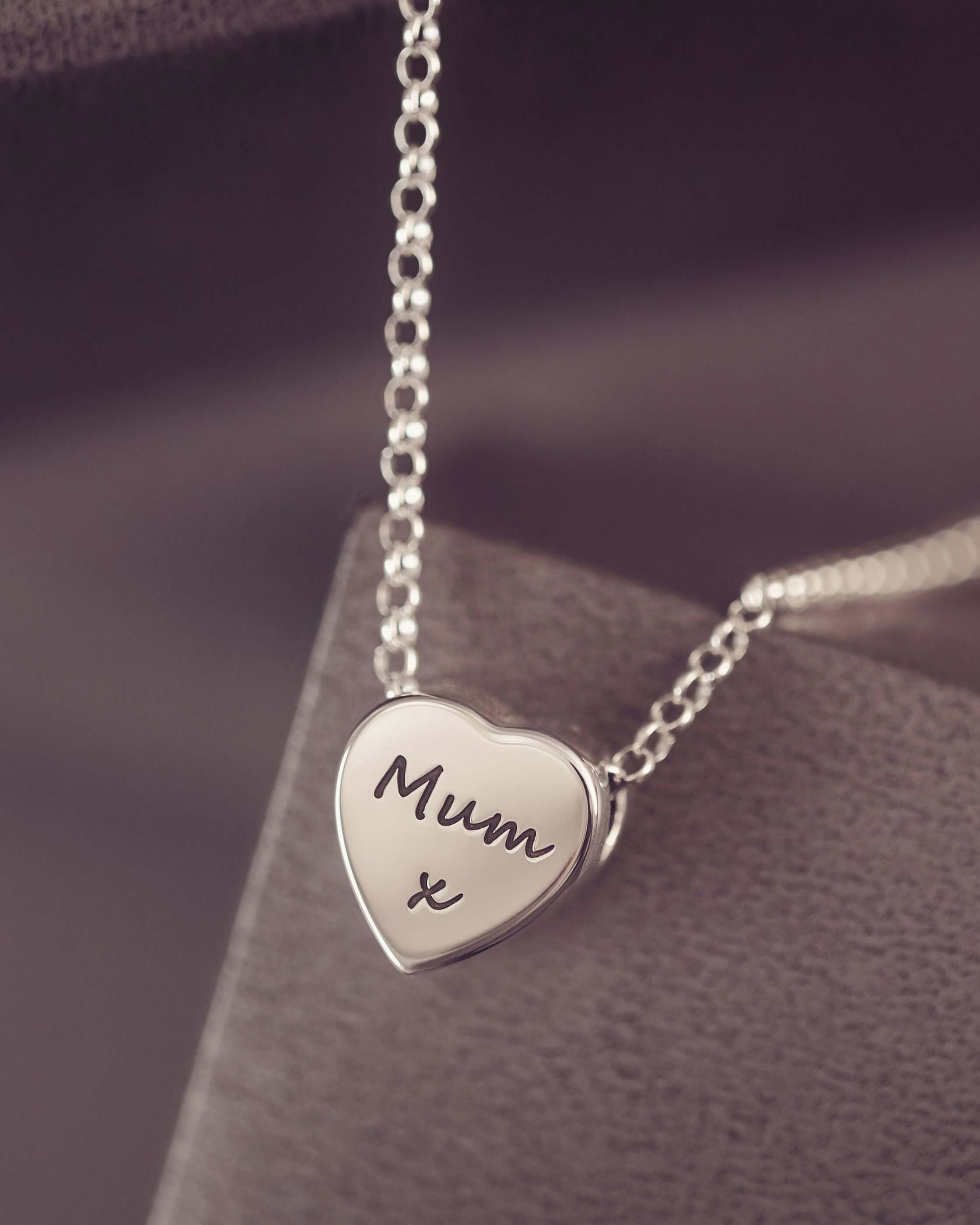 Silver heart-shaped pendant necklace with 'Mum x' engraving on a dark surface