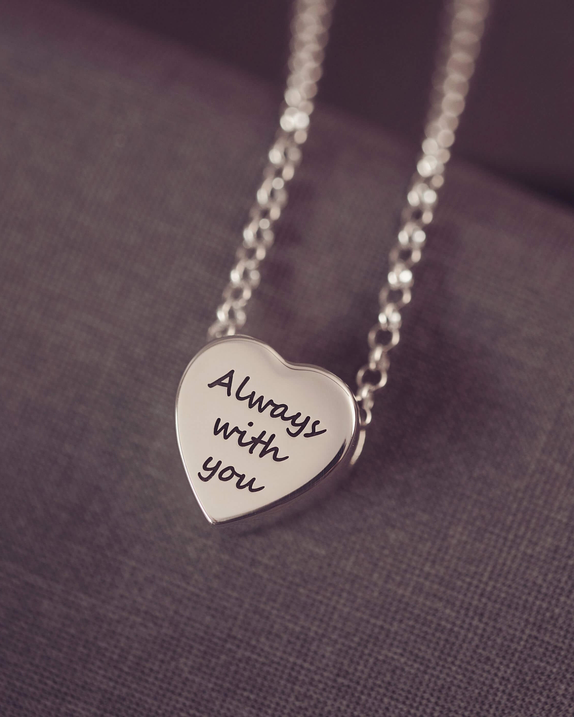 Silver heart-shaped pendant necklace with 'Always with you' engraving on a purple fabric background