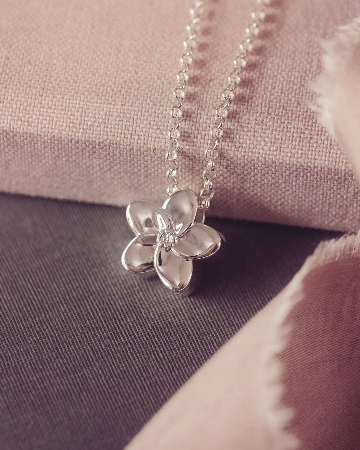 A silver flower pendant necklace resting on a textured pink fabric surface with a dark gray fabric background.