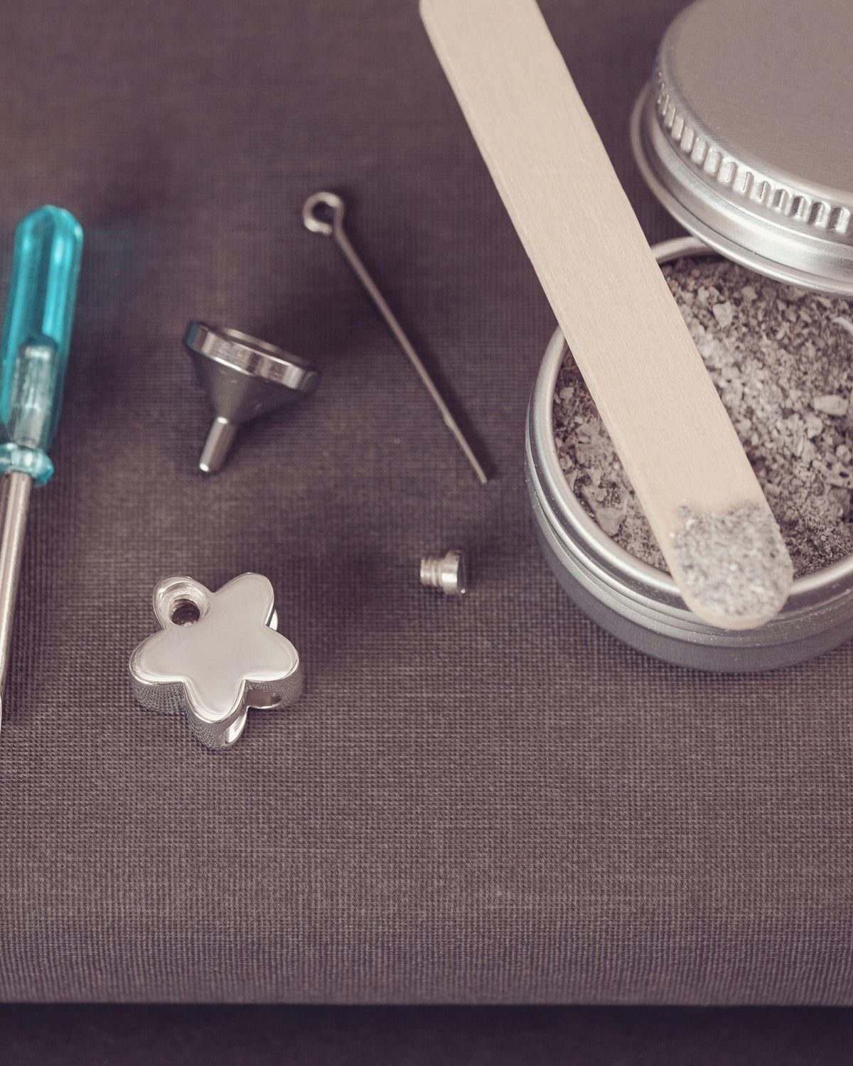 Silver jewelry-making supplies arranged on a dark textured surface, including a small turquoise-handled screwdriver, metal funnel, pin, flower-shaped charm and an open tin of ashes with a wooden spatula.