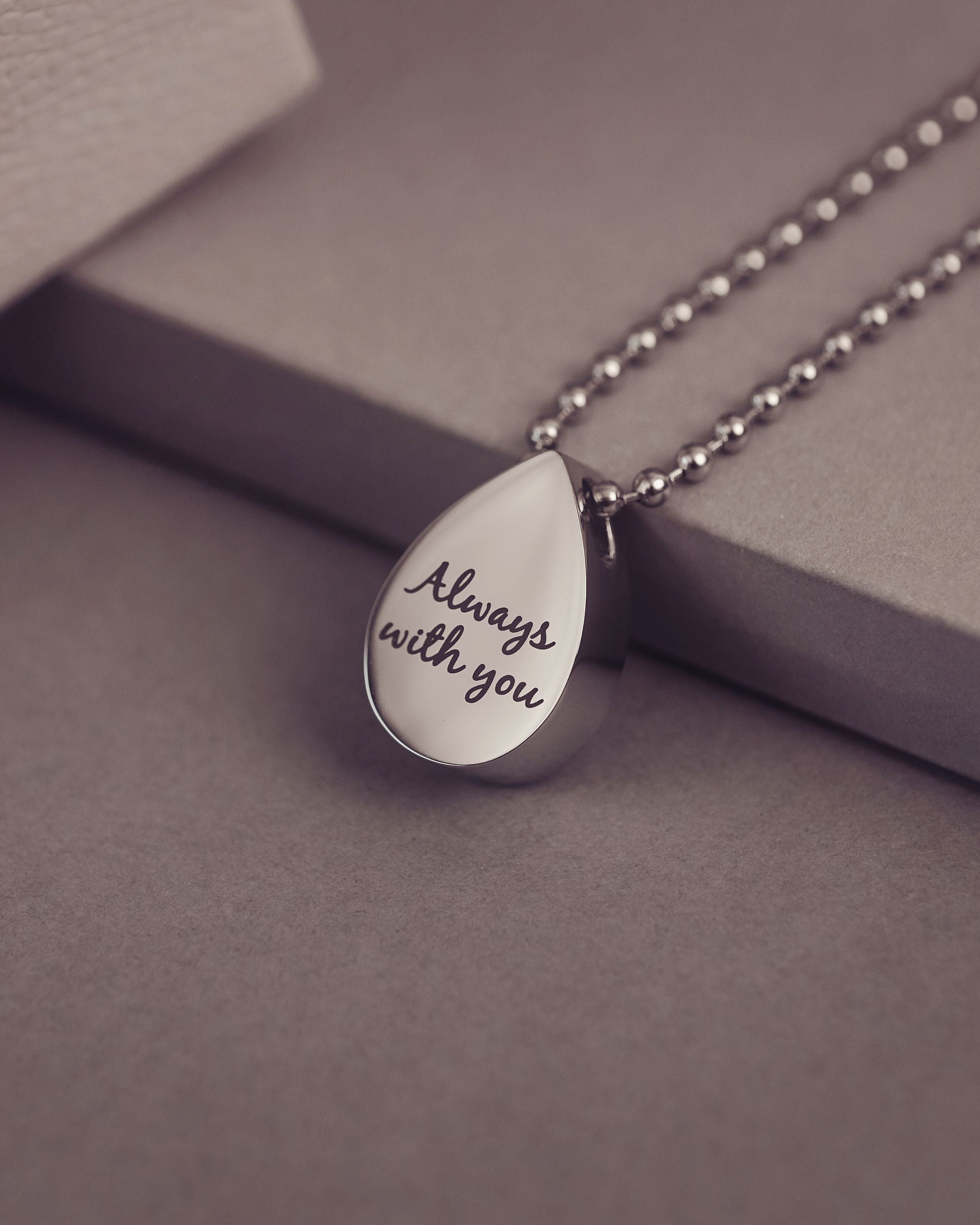 Silver teardrop pendant necklace with 'Always with you' engraving on a gray surface
