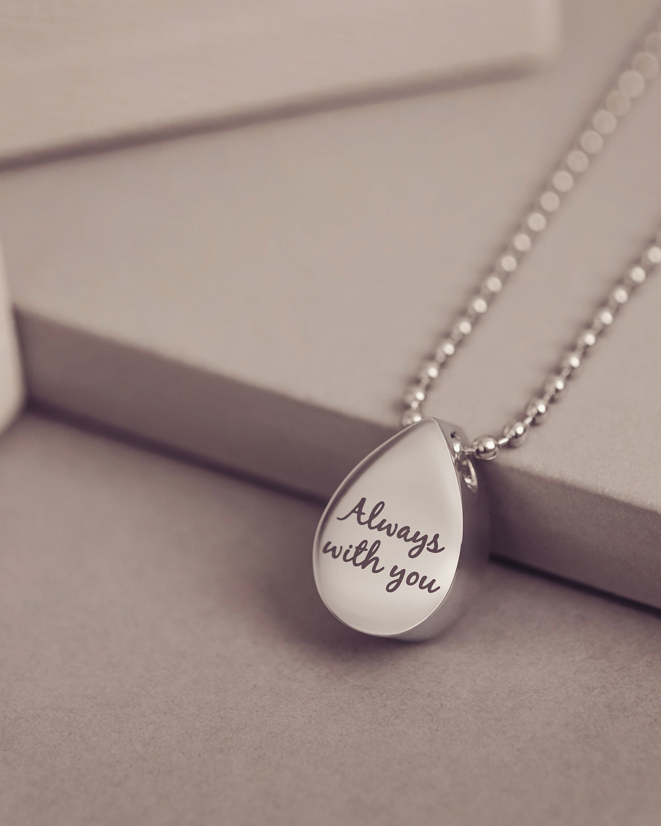 Teardrop-shaped pendant with 'Always with you' engraving on a chain, set against a neutral background.