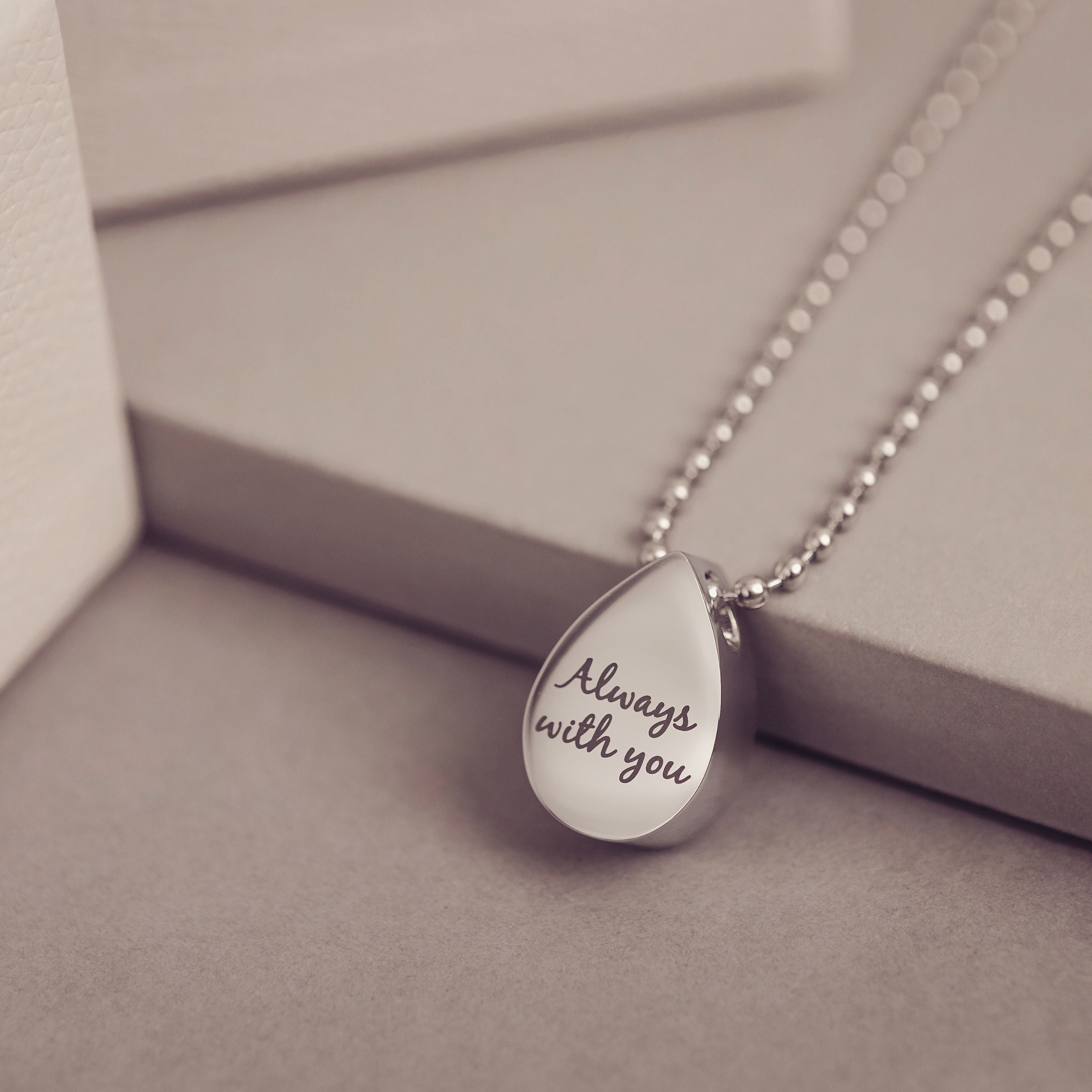 Teardrop-shaped pendant with 'Always with you' engraving on a chain, set against a neutral background.
