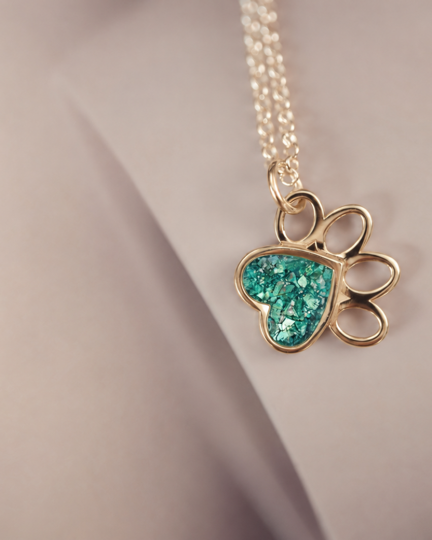 A gold paw print pendant necklace with a glittery turquoise heart inlay, resting on a neutral beige surface.
