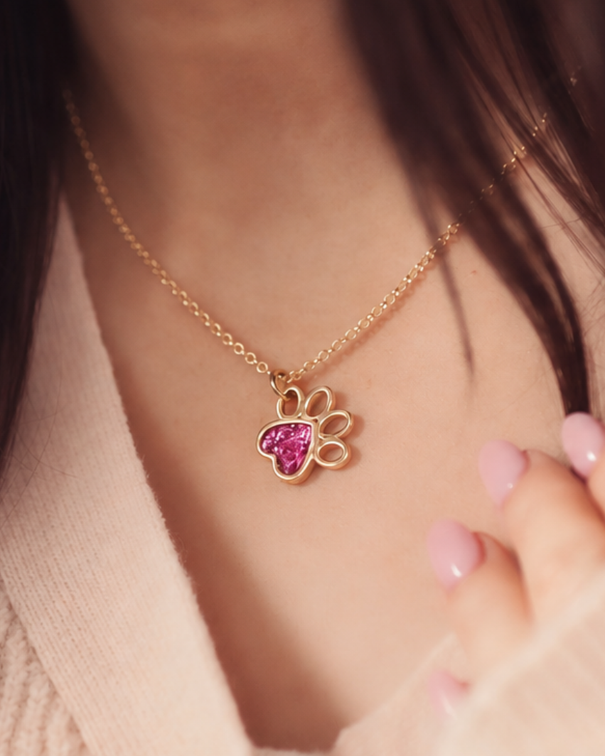A close-up lifestyle shot of a gold chain necklace with a paw print pendant featuring a pink heart-shaped center, worn by a person in a beige knit top.
