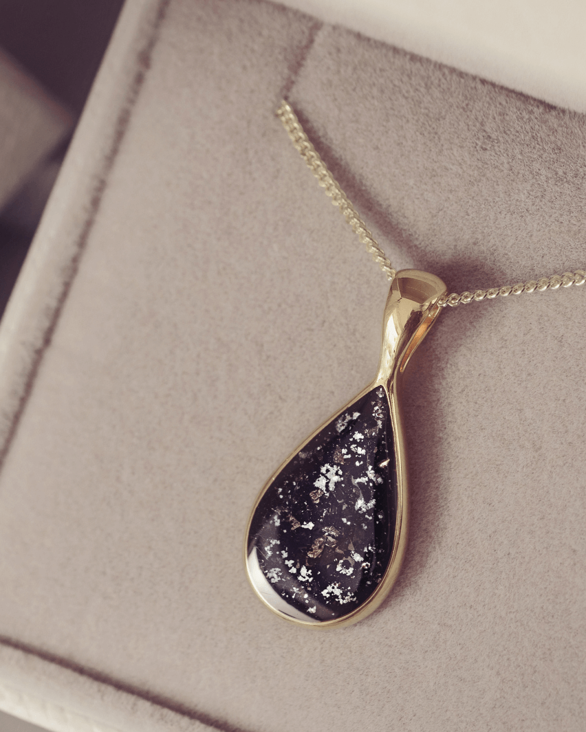 Gold chain with a teardrop-shaped pendant, including ashes in a black setting, on a soft surface