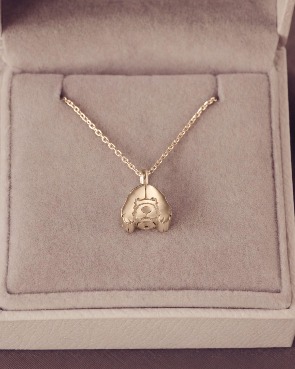 A gold pendant necklace displayed inside an open light beige jewelry box. The pendant is of a stylized dog, suspended from a delicate chain.