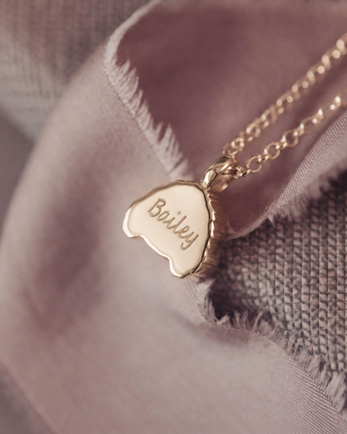 A close-up of a gold necklace with a small engraved pendant that reads 'Bailey', resting on soft dusty pink fabric and textured gray woven material.