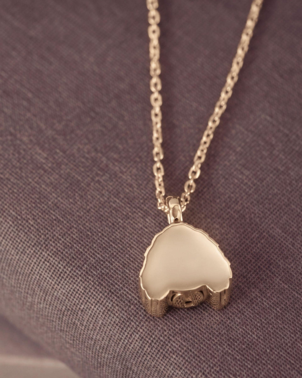 A gold pendant necklace with a smooth, polished dog-shaped pendant resting on a textured gray fabric background.