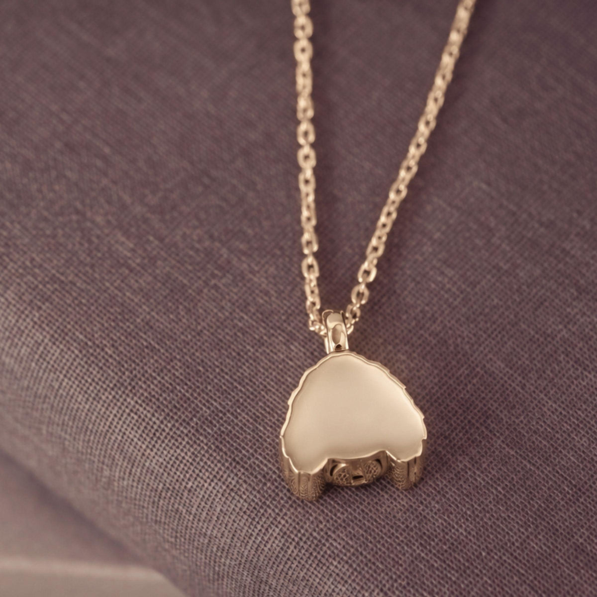 A gold pendant necklace with a smooth, polished dog-shaped pendant resting on a textured gray fabric background.