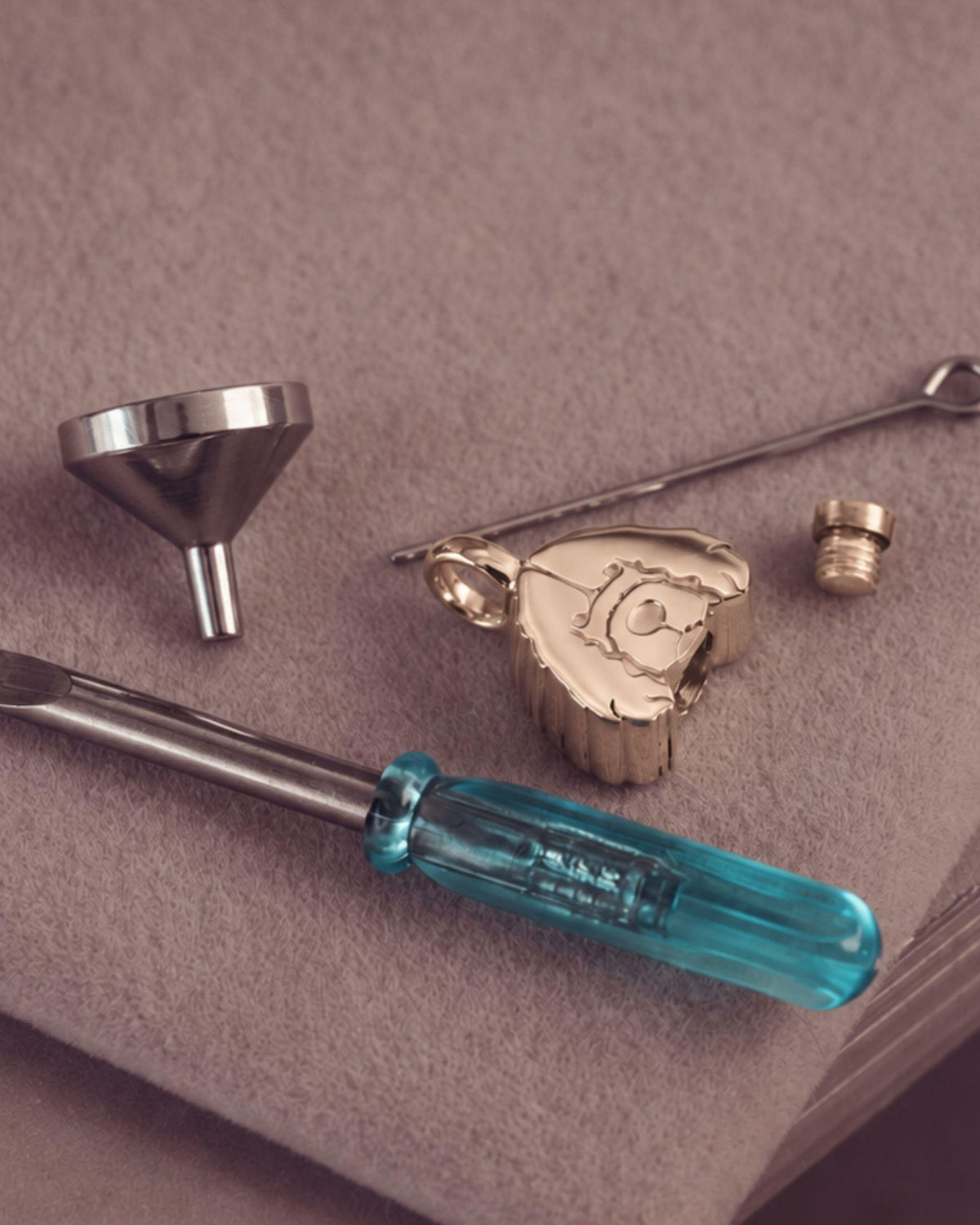 A jewelry filling kit with a dog-shaped gold pendant, a silver funnel, a small metal tool, a flat head screwdriver with a translucent blue handle. Arranged on a textured taupe surface.