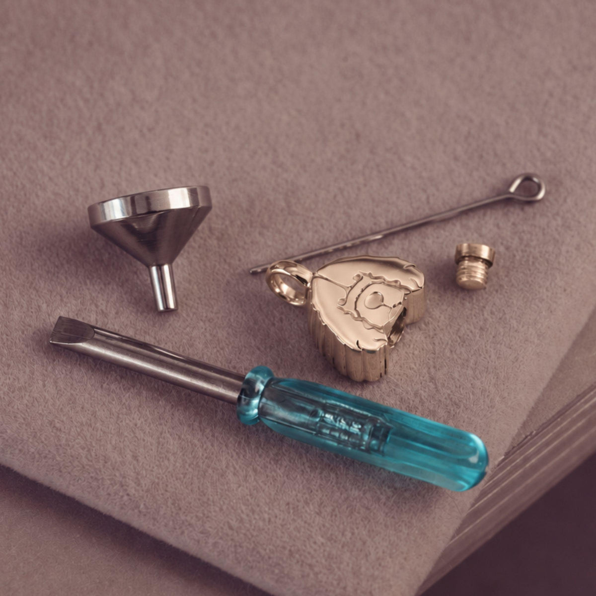 A jewelry filling kit with a dog-shaped gold pendant, a silver funnel, a small metal tool, a flat head screwdriver with a translucent blue handle. Arranged on a textured taupe surface.
