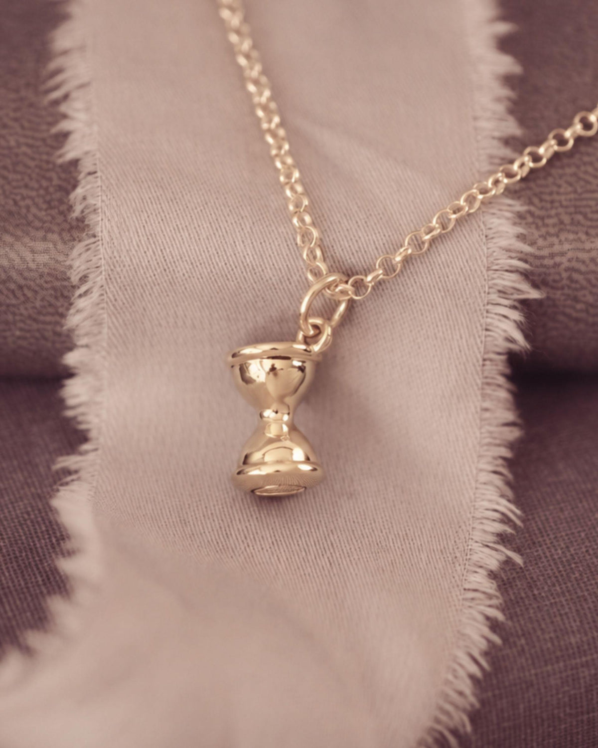 A gold-toned pendant necklace with a small hourglass charm, displayed on a textured fabric background with a frayed beige ribbon.